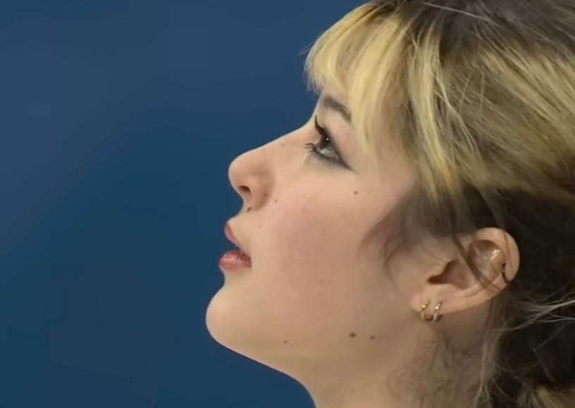 A silhouette of a female-presenting face, whose hair is striped in blonde and brown. The chin tilts skyward with a blue backdrop. Gold, silver, and dark metal hoop earrings line up on the l