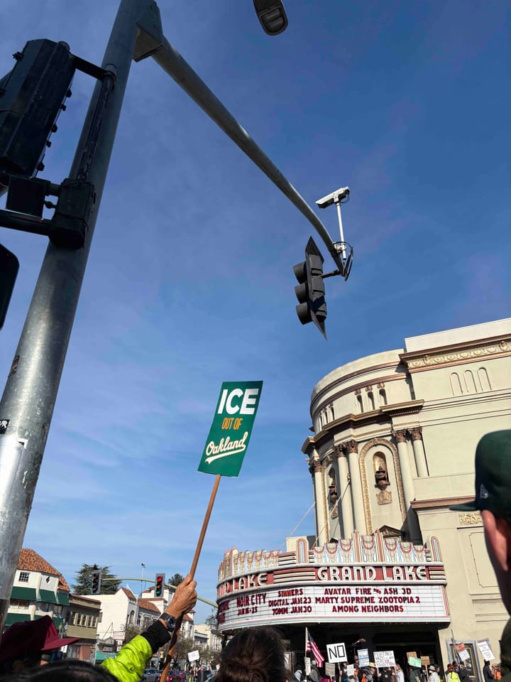 A green sign with the words, "ICE Out of Oakland" features a city name in Athletics font. Throngs of protestors gather at the landmark Grand Lake Theatre in view of Flock camera.
